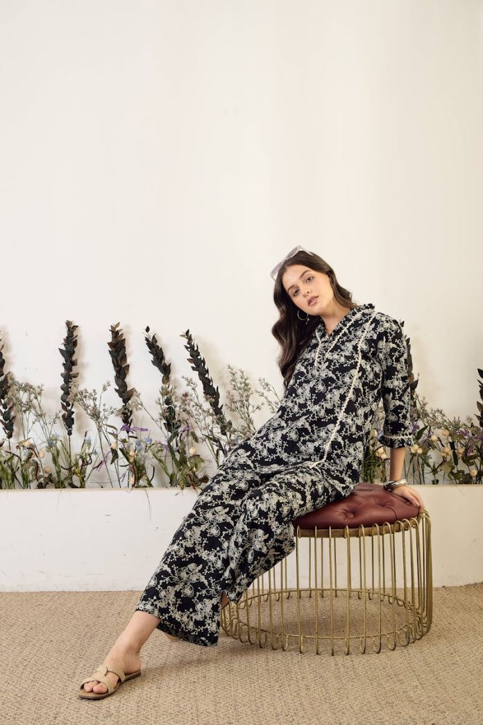 Woman in floral jumpsuit sitting on stool.