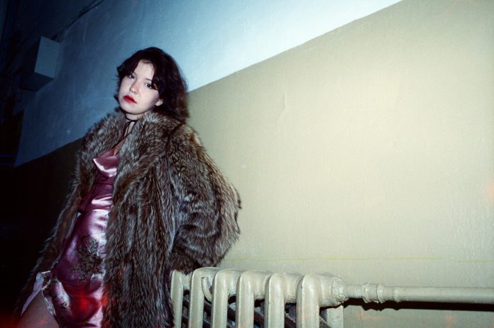 Woman in fur coat and shiny dress stands by radiator.